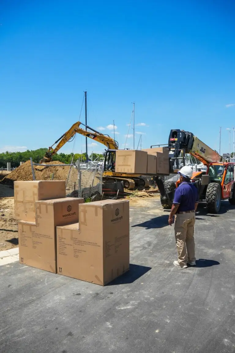 A construction supervisor in a hard hat watches a telehandler lift Woodcraft Hospitality furniture boxes at a waterfront building site, with an excavator and marina sailboats in the background.